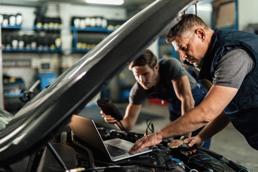 Zwei Männer in Arbeitskleidung arbeiten an einem Auto in einer Werkstatt.