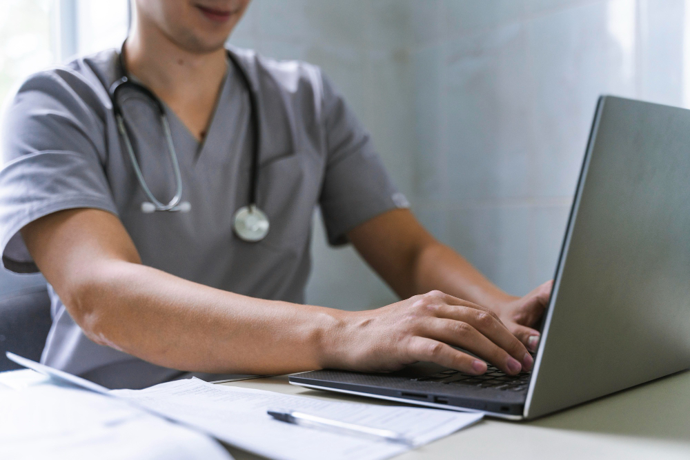 Foto einer Person mit einem Stethoskop um den Hals und einem kurzärmligen Arztkittel arbeitet am Laptop.