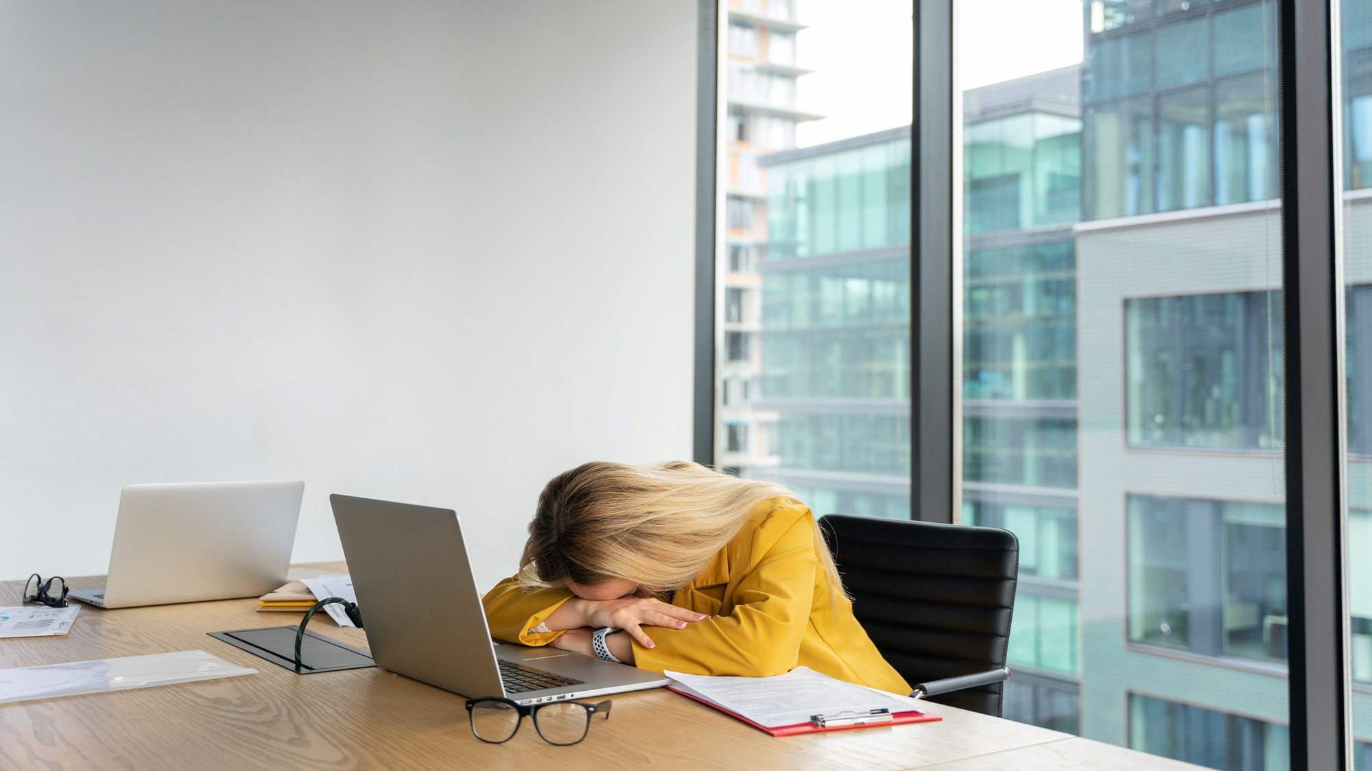 Foto einer Frau in einem Büro. Ihren Kopf hat sie auf dem Tisch vor ihrem Laptop abgelegt hat.