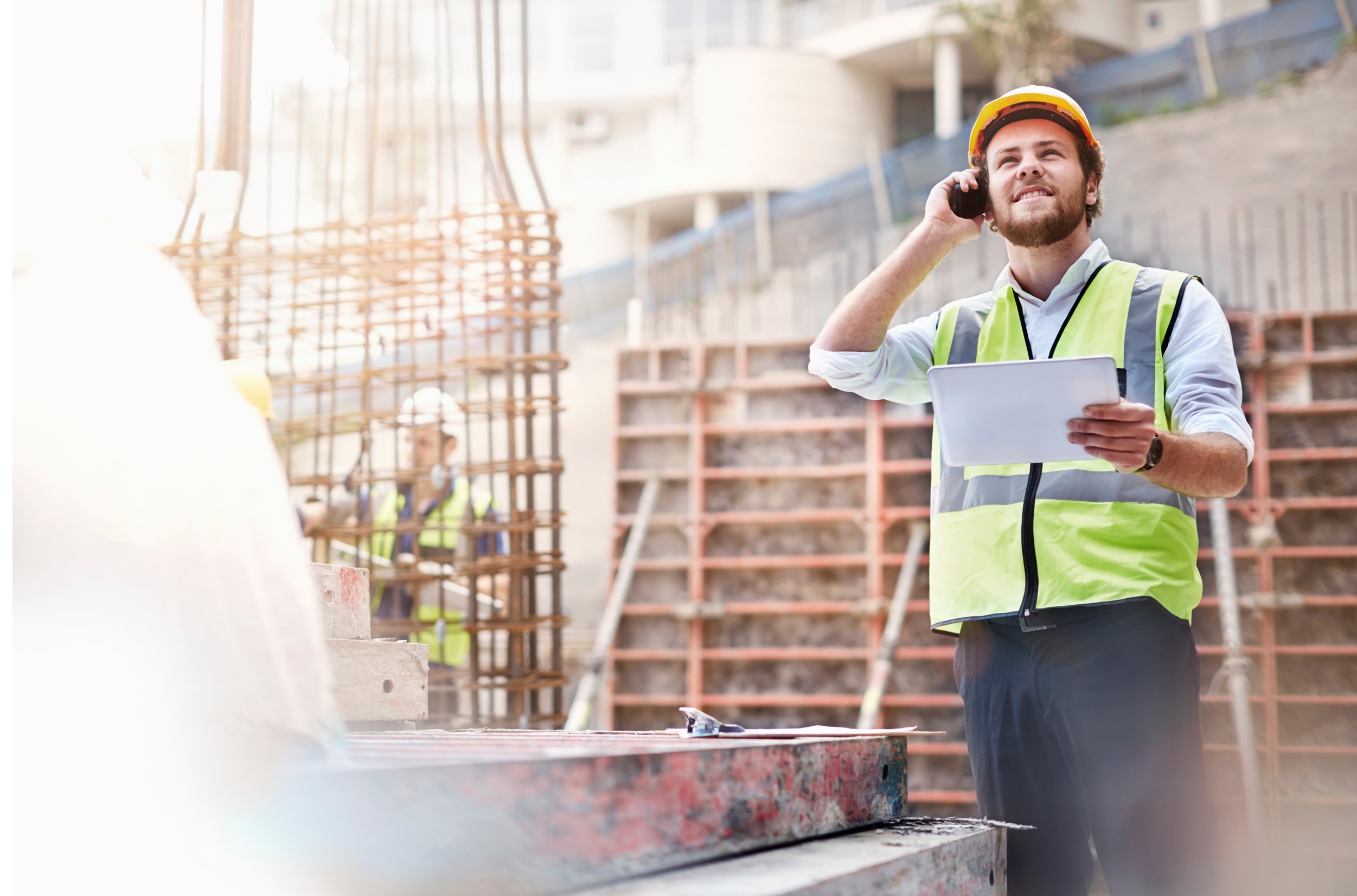 Foto eines Mannes, der auf einer Baustelle steht mit einer Warnweste und einem Schutzhelm. Er telefoniert und hält ein Ipad in der Hand.